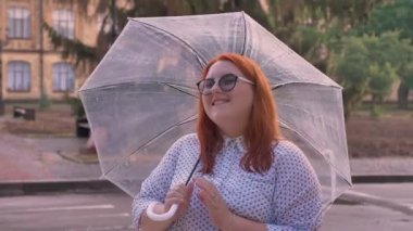 Gözlüklü şişman zencefil kız yağmurlu havalarda, dönüyor şemsiye, kameraya, gülümseyen, kavram rüya izlerken duruyor