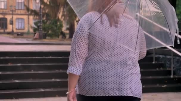 Fat ginger girl with glasses is alking up little stairs in park in rainy weather, holding umbrella — Stock Video