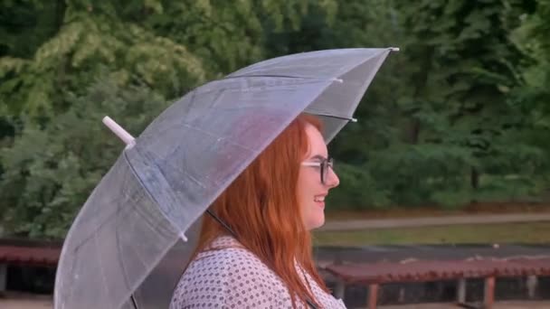 Fet ingefära tjej med glasögon passerar på väg i park i regnigt väder, kommer till byggnaden, innehar paraply — Stockvideo