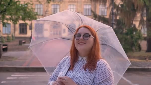 Gordura gengibre menina com óculos está de pé no tempo chuvoso, spining guarda-chuva, assistindo a câmera, sorrindo, construindo no fundo — Vídeo de Stock