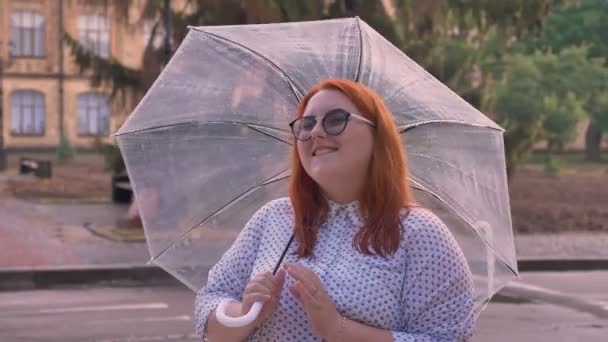 Gros gingembre fille avec des lunettes est debout par temps pluvieux, parapluie filant, regarder à la caméra, sourire, concept de rêve — Video