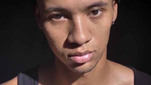 Jovem homem africano bonito com piercing na orelha olhando para a câmera com expressão séria, fundo preto — Vídeo de Stock