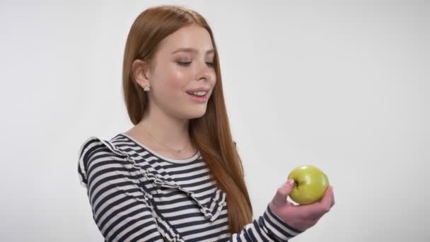 Chica jengibre dulce joven está mordiendo manzana, ofrécelo, concepto de comunicación, fondo blanco — Vídeos de Stock