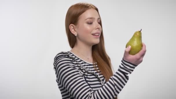 Молодая милая рыжая девушка кусает грушу, наслаждаясь им, белый фон — стоковое видео