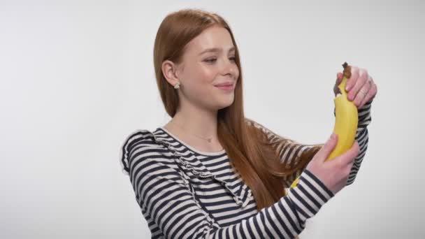 Young sweet ginger girl is peeling banana, biting it, enjoying, snack concept, white background — Stock Video
