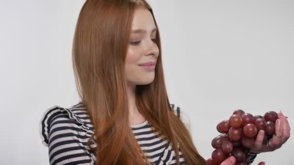 Jeune fille au gingembre doux tient le raisin, offrant, regarder à la caméra, fond blanc — Video