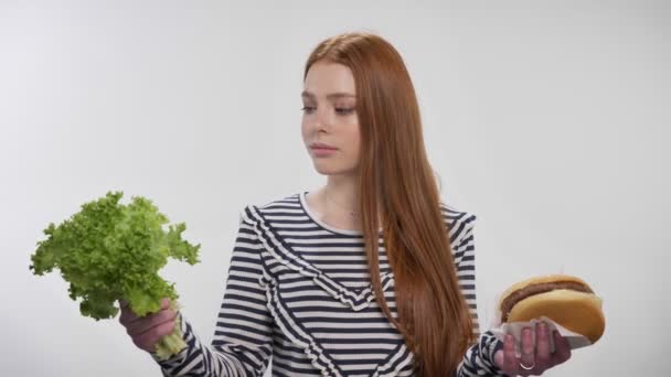 Молодая милая рыжая девушка выбирает между салатом и бургером, зеленый выбор, белый фон — стоковое видео