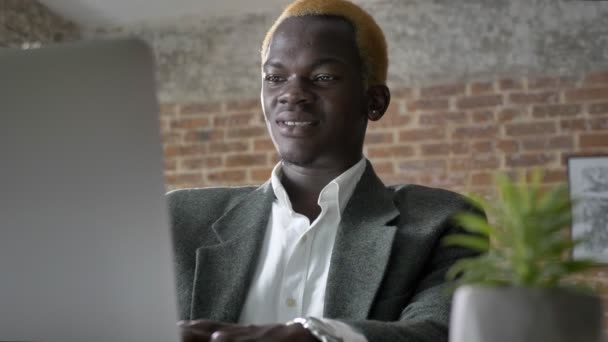Junge afrikanische blonde Geschäftsmann tippt auf Laptop und sitzt in modernem Büro, zuversichtlich, lachend — Stockvideo
