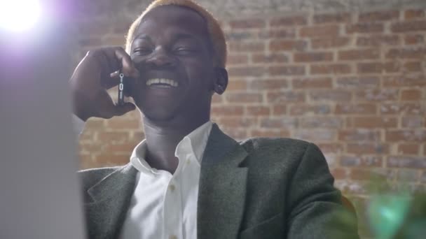 Giovane africano felice uomo d'affari bionda parlando al telefono e seduto in ufficio moderno, ridendo e sorridendo — Video Stock