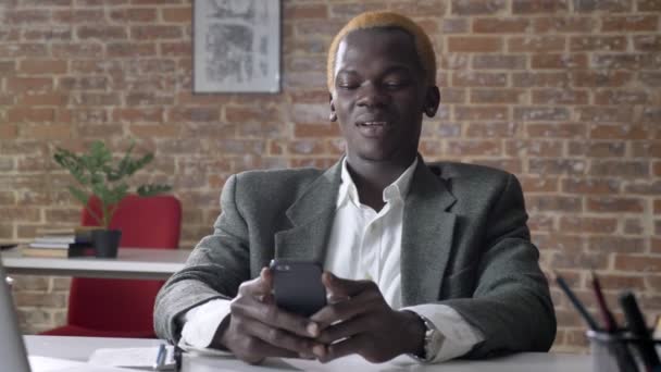 Молодий африканський модний бізнесмен з світлим волоссям друкує по телефону і сидить в офісі, чоловік з проколотим вухом — стокове відео