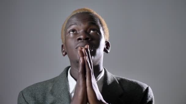 Молодой африканский блондин в костюме, держась за руки и молясь, изолированный на сером фоне — стоковое видео