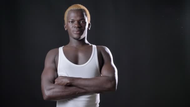 Unga afrikanska blonda mannen i vit skjorta står med korsade händer och tittar på kameran, självsäker och allvarliga, svart studio bakgrund — Stockvideo