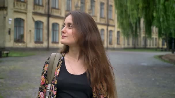 Молода чарівна жінка, дивлячись навколо і стоячи в парку біля коледжу, щаслива і весела — стокове відео