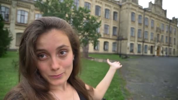 若くてきれいな女性ブロガーのブログやビデオ チャットを撮影、建物、笑みを浮かべて大学近くの公園で立っています。 — ストック動画