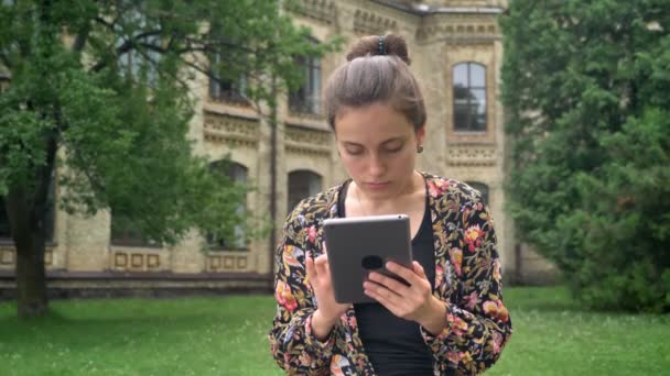 Mujer bonita joven en chaqueta de colores navegando en la tableta y sentado en el parque cerca del campus universitario, encantador y atractivo — Vídeo de stock