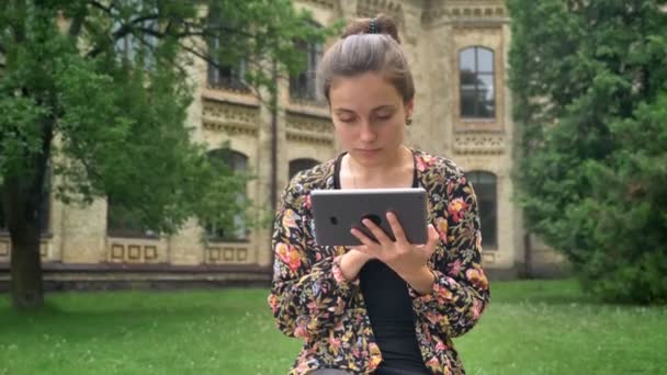 Young beautiful woman typing on tablet and sitting on bench near university in park with grass, smiling at camera — Stock Video