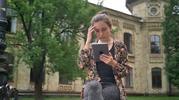 Unga vackra kvinnliga högskolestudent holding tablett och sitta på bänken i parken nära university — Stockvideo