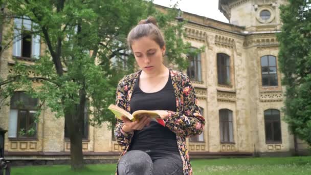 Joven hermosa mujer sentada y leyendo libro, saludando a alguien y marchándose, parque cerca de antecedentes universitarios — Vídeos de Stock