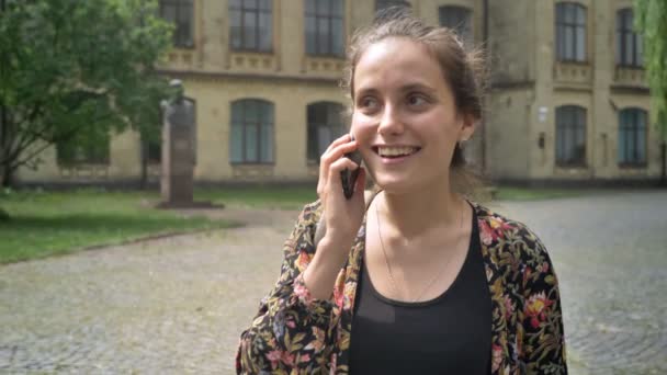 Joven mujer alegre caminando por la calle cerca de la universidad y hablando por teléfono, encantadora estudiante universitaria — Vídeos de Stock