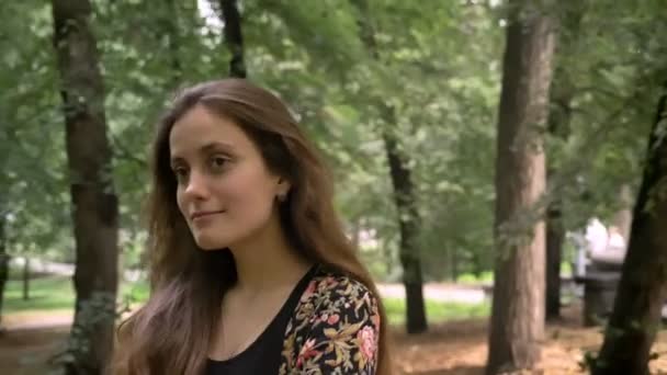 Giovane bella donna con i capelli lunghi passeggiando nel parco con alberi verdi e sorridenti, felici e allegri — Video Stock