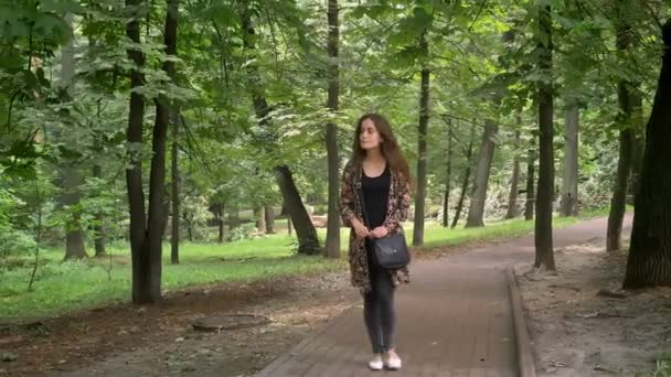 Mujer encantadora joven en chaqueta de colores caminando en el parque con árboles verdes y mirando a su alrededor, feliz y alegre — Vídeos de Stock