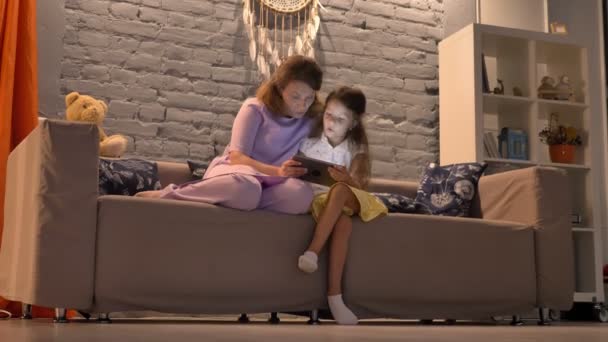 Mère et sa petite fille utilisant la tablette, assis ensemble sur le canapé dans le salon moderne, concept de famille avec gadget — Video