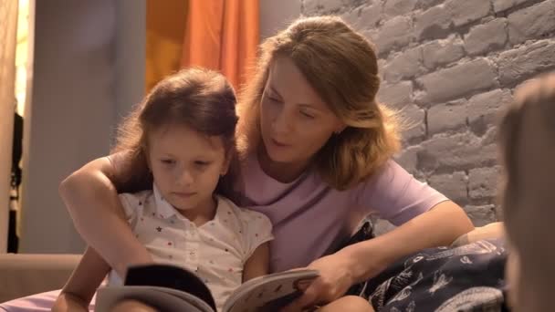 A kislány, anya és a gyerek ül össze a kanapé, modern nappalival, családi koncepció, bent a szülő olvasókönyv — Stock videók
