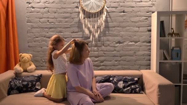 Kleine Tochter bürstet der Mutter die Haare mit der Haarbürste, sitzt auf dem Sofa im modernen Wohnzimmer, Familienkonzept drinnen — Stockvideo