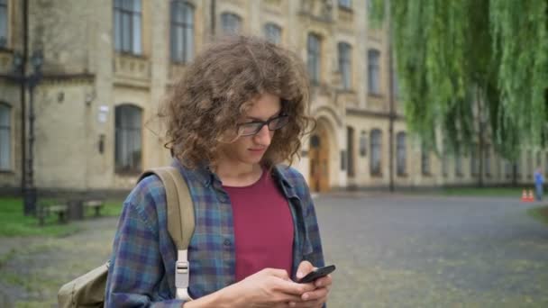 Młody przystojny student z kręconymi włosami w okulary wiadomości tekstowe na telefon i trzyma plecak, stojąc w parku, w pobliżu Uniwersytetu — Wideo stockowe