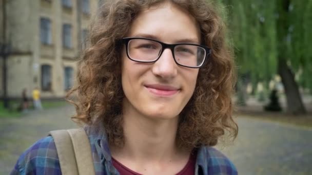 Jovem homem bonito em óculos com cabelo encaracolado sorrindo para a câmera e de pé na rua perto do prédio da faculdade, feliz — Vídeo de Stock