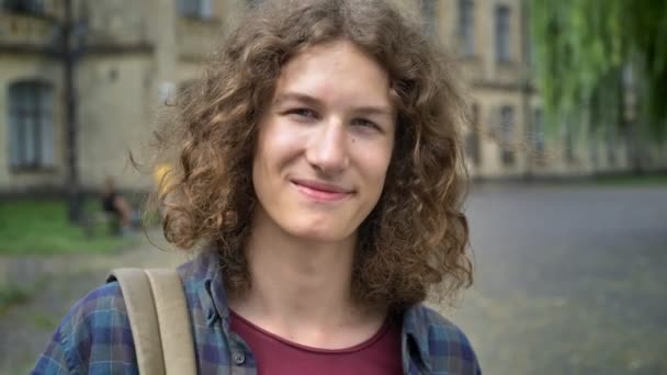 Jeune homme beau avec de longs cheveux bouclés regardant la caméra et souriant, tenant sac à dos et debout dans la rue près de l'université — Video