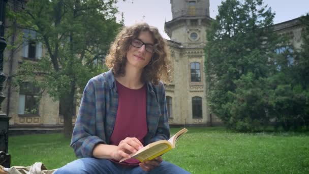 Молодий красивий студент в окулярах з кучерявим волоссям читає книгу і дивиться на камеру, сидячи в парку біля будівлі університету — стокове відео