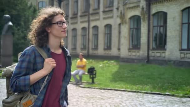 Chytrý hloupý student s vlnité vlasy v brýlích chodit na vysokou, drží batoh na jednom rameni, dotýká vlasů — Stock video