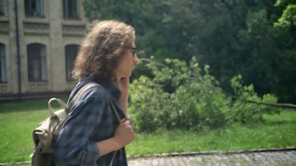 Jonge knappe man met krullend lang haar in bril opgelopen naar de Universiteit en praten over telefoon, straat in de buurt van college achtergrond — Stockvideo