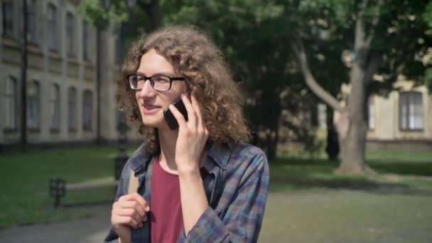 Jonge gelukkig student met krullend haar gaan naar school en praten over telefoon, wandelen in het park in de buurt van Universiteit, overdag — Stockvideo