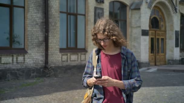 Mladý pohledný student s batohem odcházely z college a textové zprávy na telefonu, chůzi na ulici univerzitní budovy — Stock video