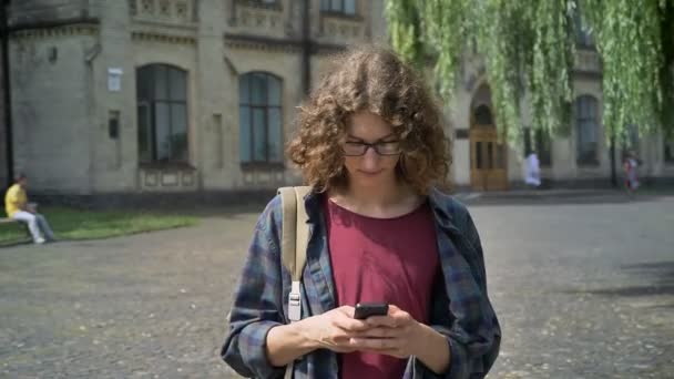Giovane studente nerd con lunghi capelli ricci a piedi e digitando al telefono, parco vicino all'università o strada vicino allo sfondo del college — Video Stock