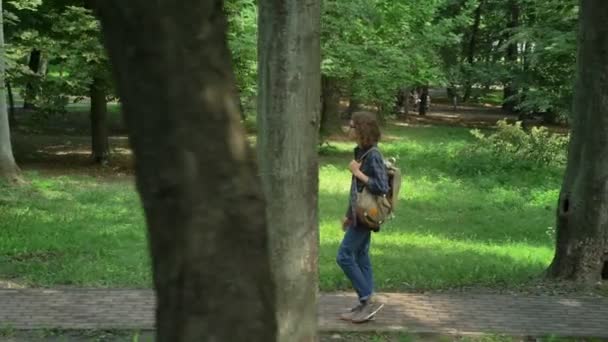 Mahasiswa kutu buku dengan rambut keriting panjang berjalan di taman dan melihat ke depan, pohon dan rumput di sekitar, sinar matahari di luar ruangan — Stok Video