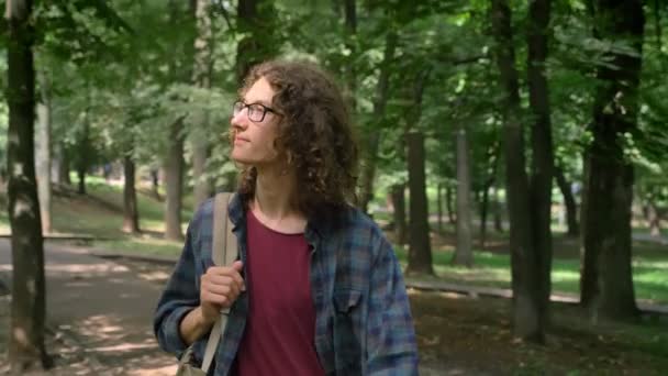 Nerdy mladý student v brýlích s batohem procházky v parku a rozhlédl se kolem sebe, slunce svítí jasně venku — Stock video