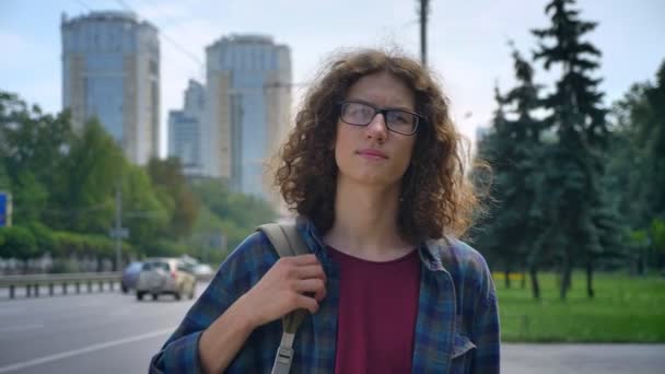 Giovane studente nerd in occhiali con lunghi capelli ricci in attesa di taxi, in piedi alla fermata dell'autobus e guardando avanti, sfondo urbano della città — Video Stock
