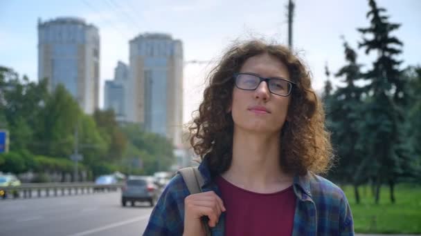 Jovem nerd de óculos com cabelos longos encaracolados esperando por táxi ou ônibus, olhando para a frente e de pé na rua perto da estrada, durante o dia — Vídeo de Stock