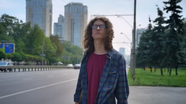 Smart young man in glasses with long curly hair waiting for taxi and touching his hair, standing on bus stop near road in urban city — Stock Video