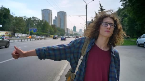 Jeune étudiant charmant dans des lunettes avec de longs cheveux ondulés en attente de taxi et debout près de la route avec des voitures, fond urbain moderne de la ville — Video