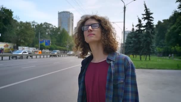 Homem bonito jovem em óculos com cabelo ondulado longo esperando por táxi ou ônibus e olhando para a frente, de pé na rua urbana perto da estrada — Vídeo de Stock