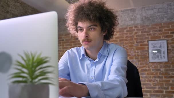 Programador nerd engraçado com codificação de cabelo encaracolado no laptop e sentado à mesa no escritório moderno — Vídeo de Stock