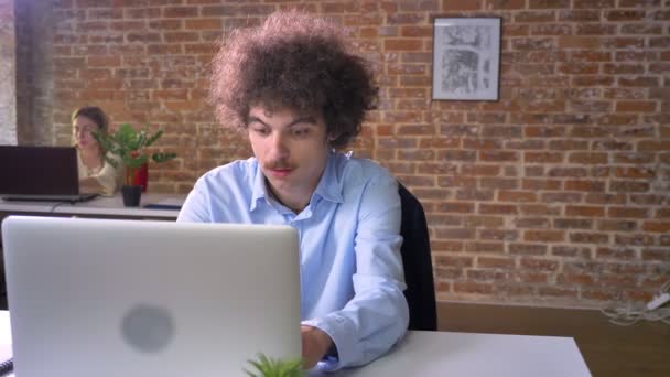 Смішний ботанік програміст з вусами і кучерявим кодуванням волосся на ноутбуці, сидячи за столом, сучасний офісний фон — стокове відео