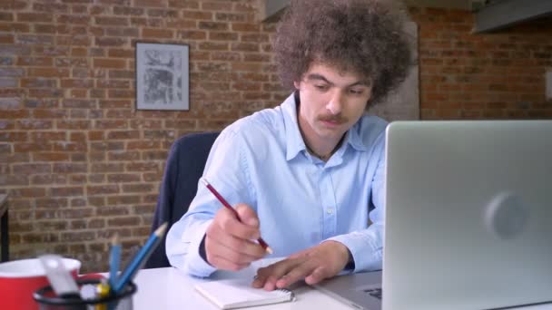 Молодой ботаник-бизнесмен пишет заметки с ноутбука, сидит в современном офисе, серьезный и сосредоточенный — стоковое видео