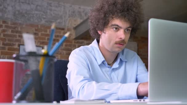 Empresario nerd serio con bigote y pelo rizado trabajando en el ordenador portátil y tomando notas, sentado en la oficina moderna — Vídeos de Stock