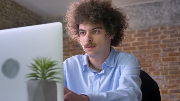 Homem nerd engraçado com cabelo encaracolado volume pensando e digitando no laptop, sentado no escritório moderno, concentrado e determinado — Vídeo de Stock