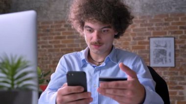 Genç adam kıvırcık saçlı ve kredi kartı kullanarak ve modern ofis masada oturan telefon tutarak bıyık konsantre